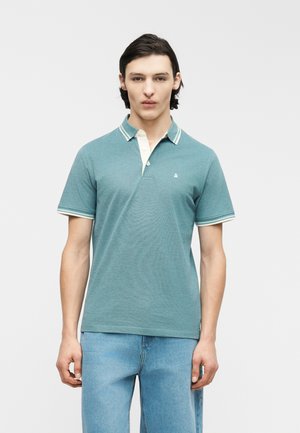 Jonge man met donker haar, gekleed in een teal polo met korte mouwen en witte bies, en lichte blauwe jeans, staand tegen een effen achtergrond.