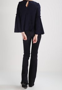 Blusa blu navy con maniche a campana, dettaglio di apertura a chiave sul retro, abbinata a jeans neri a zampa d'elefante e scarpe in vernice. Tessuto dalla consistenza liscia.