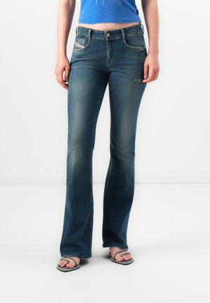 Mujer vestida con jeans bootcut azul oscuro, top corto azul y sandalias negras de punta abierta con tiras finas, de pie contra un fondo blanco.