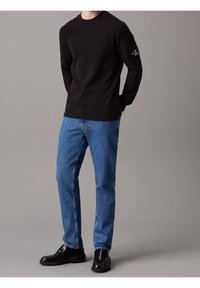 Black long-sleeve sweater with round neck, textured fabric, displaying a small logo patch. Paired with blue straight-leg jeans and black loafers.