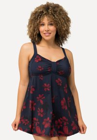 Femme aux cheveux bouclés portant une robe de bain bleu marine avec un motif floral rouge, tenant l'ourlet et souriant devant un fond uni.