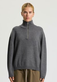 Donna che indossa un maglione a collo alto grigio a costine con una zip frontale e pantaloni marroni, su uno sfondo chiaro e uniforme.