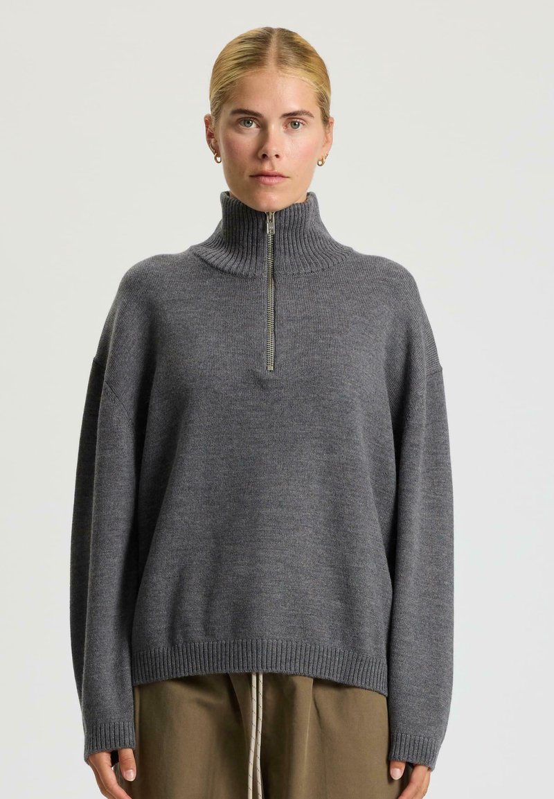 Donna che indossa un maglione a collo alto grigio a costine con una zip frontale e pantaloni marroni, su uno sfondo chiaro e uniforme.
