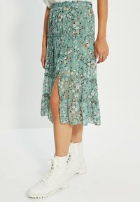Jupe maxi fleurie en vert avec des niveaux superposés et volantés. Présente une fente sur le côté et une taille élastique. Portez-la avec des bottes blanches à lacets.