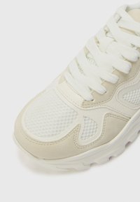 Beige treningsko laget av mesh og suede-materiale, tykke hvite snøring, og en polstret såle med en strukturert gummigrep.