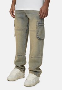 Jasnoniebieskie spodnie cargo z denimu o wyblakłym wykończeniu; posiadają dwie boczne kieszenie i szczegółowe przeszycia. Noszone z białymi sneakersami.