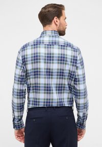 Camicia a maniche lunghe a quadri in blu chiaro, blu navy e verde. Presenta un colletto con bottoni e polsini regolabili. Indossata con pantaloni blu navy.