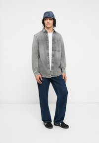Grijze denim overhemd met voorzak, in laagjes over een wit T-shirt; gecombineerd met blauw wijde jeans en zwarte sneakers met witte accenten.