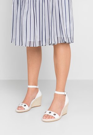 Białe sandały na koturnie z tekstylnym pokryciem na obcasa, ozdobione paskami i metalicznymi akcentami na palcach. Dopasowane do plisowanej spódnicy w paski.