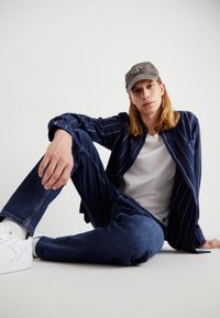 Koszula w granatowe paski na guziki, biała koszulka, niebieskie dżinsy i białe sneakersy. Czapka jest szara z napisem. Model siedzi z jedną nogą zgiętą.