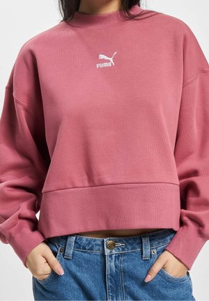 Woman wearing a cropped, dusty pink Puma sweatshirt and blue jeans, hands in pockets, cropped at mid-torso.