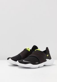 Czarne sportowe buty wsuwane z teksturowanym dzianinowym wierzchem, białą podeszwą i neonowożółtymi akcentami. Charakteryzują się smukłym, niskoprofilowym fasonem.