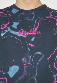 Gros plan sur une chemise foncée avec des motifs abstraits bleus et roses et un logo "ellesse" rose vif brodé sur la poitrine.
