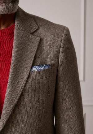 Brauner Fischgräten-Blazer mit eingekerbtem Revers, ein blaues florales Einstecktuch, getragen über einem roten Kabelstrickpullover.