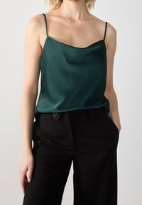 Camisola verde di raso con spalline sottili, scollatura a barca e texture liscia, abbinata a pantaloni neri a vita alta con dettagli di bottoni.