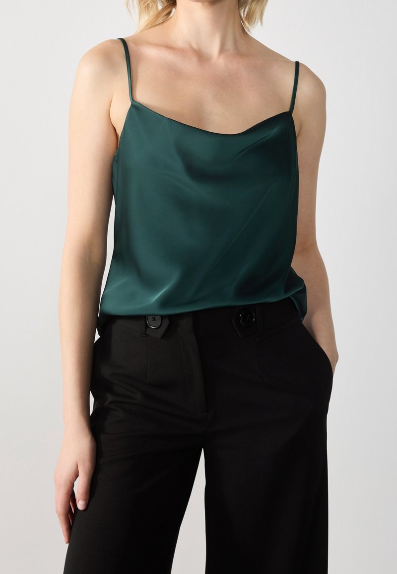 Camisola verde di raso con spalline sottili, scollatura a barca e texture liscia, abbinata a pantaloni neri a vita alta con dettagli di bottoni.