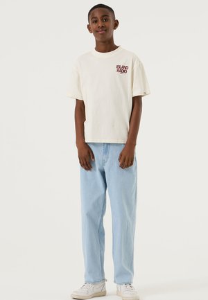 Lichtbeige katoenen T-shirt met "ISLAND RADIO" in bordeauxrood op de borst gedrukt, gecombineerd met lichtblauwe jeans met een relaxte pasvorm en witte sneakers.