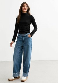 Top negro de manga larga con cuello alto combinado con jeans de mezclilla azul de pierna ancha con dobladillo deshilachado y zapatillas blancas con suela beige.