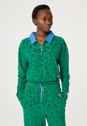 Vrouw draagt een groene, gestructureerde sweatshirt met luipaardprint, een blauwe kraag en bijpassende broek met trekkoord, handen in de zakken, kijkt opzij.