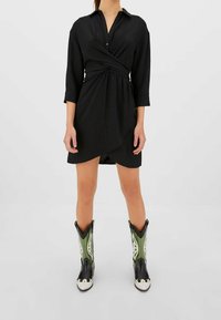 Vestido negro cruzado de manga larga, con cuello abotonado y tela texturizada, combinado con botas de vaquero verdes y negras con acentos blancos.