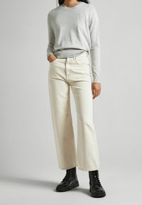 Pull gris clair associé à un jean blanc cassé à jambes larges. Le jean présente un design classique  à cinq poches. Le mannequin porte des bottes noires à lacets.