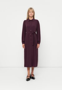Rochie lungă, de culoare prună, cu guler, închidere cu nasturi în față și un curea legată la talie. Dispune de mâneci lungi și un design drept, fluid.