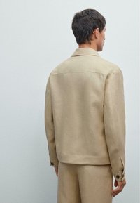 Beige Leinenjacke mit kragenförmigem Ausschnitt, Knopfmanschetten und lockerer Passform, von hinten vor einem neutralen Hintergrund gezeigt.