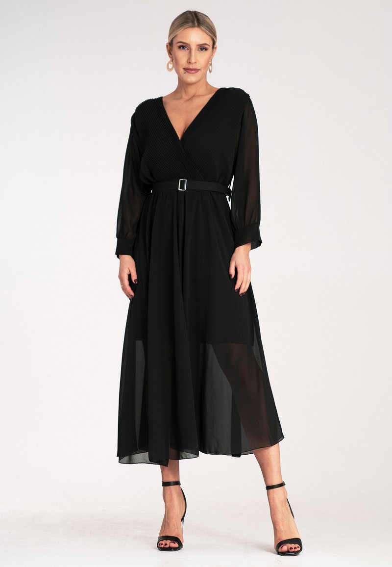 Czarna sukienka z długim rękawem w stylu dopasowanym, z teksturowaną górą, paskiem w talii i zwiewnym, przezroczystym dołem. Stylizowana z wysokimi szpilkami.