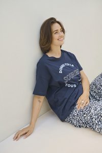 Tânără cu păr maro până la umeri stă sprijinită de un perete, zâmbind, purtând un tricou bleumarin cu text în franceză și pantaloni cu model.