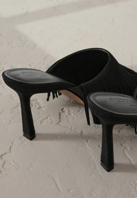 Sandales à talons hauts noires avec une finition en daim texturé, mettant en valeur des accents de franges et un design ouvert élégant. Le talon carré s'harmonise avec une esthétique stylée.