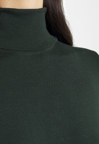 Maglione a collo alto verde scuro con una trama a coste. Il tessuto appare morbido e aderente attorno al collo e alle spalle.