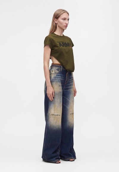 Γυναίκα που φοράει crop top σε ελαιί πράσινο με κείμενο "ARMY" και ψηλόμεσο, φαρδύ τζιν σε ξεθωριασμένο μπλε χρώμα με φθαρμένες περιοχές.