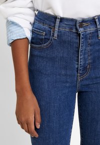 Los jeans de talle alto azul oscuro cuentan con cinco bolsillos, costuras en contraste y una textura de mezclilla suave. Una camisa blanca con puños muestra un detalle a rayas.