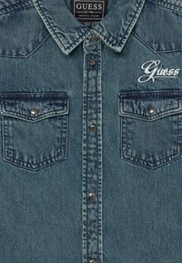 Τζιν πουκάμισο σε ανοιχτό μπλε με καμπυλωτό γιακά, κουμπιά με κλιπ, δύο μπροστινές τσέπες και κεντημένο λογότυπο "Guess" στην αριστερή πλευρά.