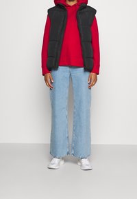 Zwarte puffer vest over een rode hoodie, gecombineerd met een lichte blauwe brede jeans en witte sneakers. Eenvoudig ontwerp met een casual fit.