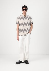 Korte mouwen shirt in roomkleur met zwart zigzagpatroon, gecombineerd met witte broek en zwarte schoenen. Het model draagt donkere zonnebril.