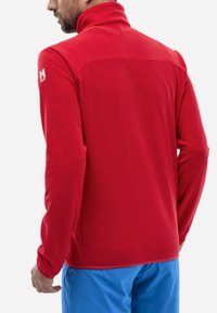 Veste polaire rouge avec un col montant, un motif texturé et des manches longues ajustées. Présente un logo sur l'épaule gauche. Assortie à un bas bleu.