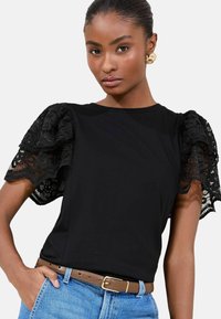 Black t-shirt with intricate lace puff sleeves, featuring a round neckline. Paired with light blue denim and a brown belt.
