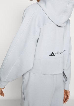 Osoba nosząca jasnoszary krótki kaptur i dopasowane spodnie dresowe z widoczną kieszenią i czarnym logo na plecach bluzy.