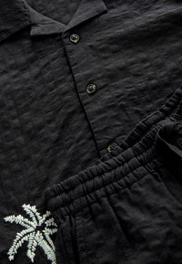 Schwarzes Hemd und passender Shorts-Satz aus strukturiertem Stoff. Das Hemd hat ein Karomuster und eine Palme-Stickerei. Knöpfe am Hemd.