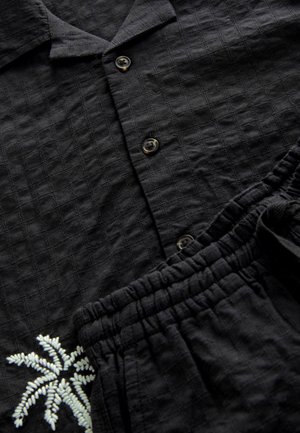 Zwarte shirt en bijpassende shortset gemaakt van geweven stof. Shirt heeft een ruitenpatroon en palmboompje borduursel. Knopen op het shirt.