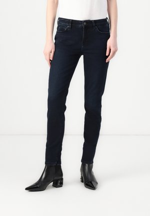 Person trägt eng anliegende dunkelblaue Jeans und spitze schwarze Stiefeletten mit Blockabsatz vor einem einfarbigen weißen Hintergrund.