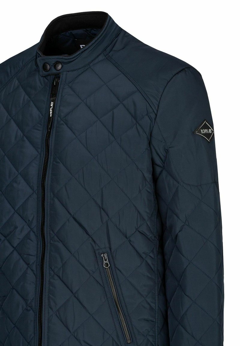 Giacca trapuntata blu navy con colletto alto, chiusura con zip e tasca laterale, caratterizzata da un logo a diamante nero sulla manica.