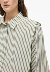 Moteris dėvi smėlio spalvos ir juodą vertikalių dryžių marškinius su struktūruotais paminkšlintais pečiais ir sidabrinėmis hoop auskarėmis.