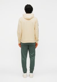 Beige hoodie met een trekkoord en geribbelde manchetten, gecombineerd met groene joggingbroek. De outfit heeft een ontspannen pasvorm en een zachte textuur.