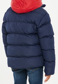 Enfant portant une doudoune bleu marine avec une capuche rouge et un jean bleu, vu de dos sur fond blanc uni.