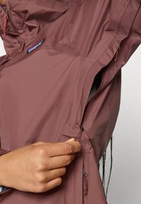 Hand die rits aanpassen van de mauve lichtgewicht jas bij de ventilatieopening onder de oksel, met het Patagonia logo patch.