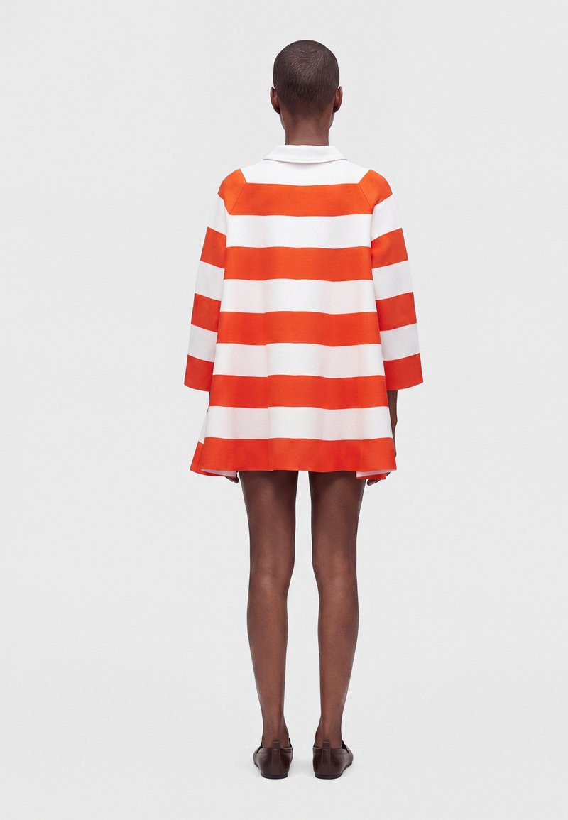 Personne aux cheveux courts portant une robe à manches longues avec de larges rayures horizontales orange et blanches, vue de dos.