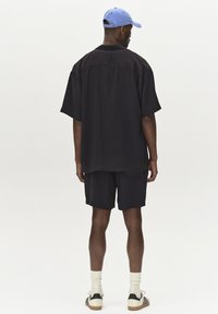 Schwarzes Oversized-Hemd mit kurzen Ärmeln, kombiniert mit passenden Shorts. Eine blaue Cap, weiße Socken und schwarz-weiße Sneakers vervollständigen das Outfit.
