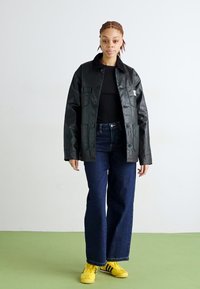 Giubbotto in ecopelle nera e lucida con due tasche frontali, abbinato a jeans blu scuro e sneakers gialle con strisce nere.